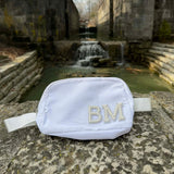 White fanny pack with 'BM' monogram on a stone surface with a water feature in the background