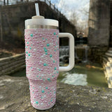 Pink beaded tumbler with a white handle and straw lid on a stone ledge with a natural background.