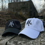 Black and white 'Mr.' and 'Mrs.' hats on a stone surface with a scenic background.