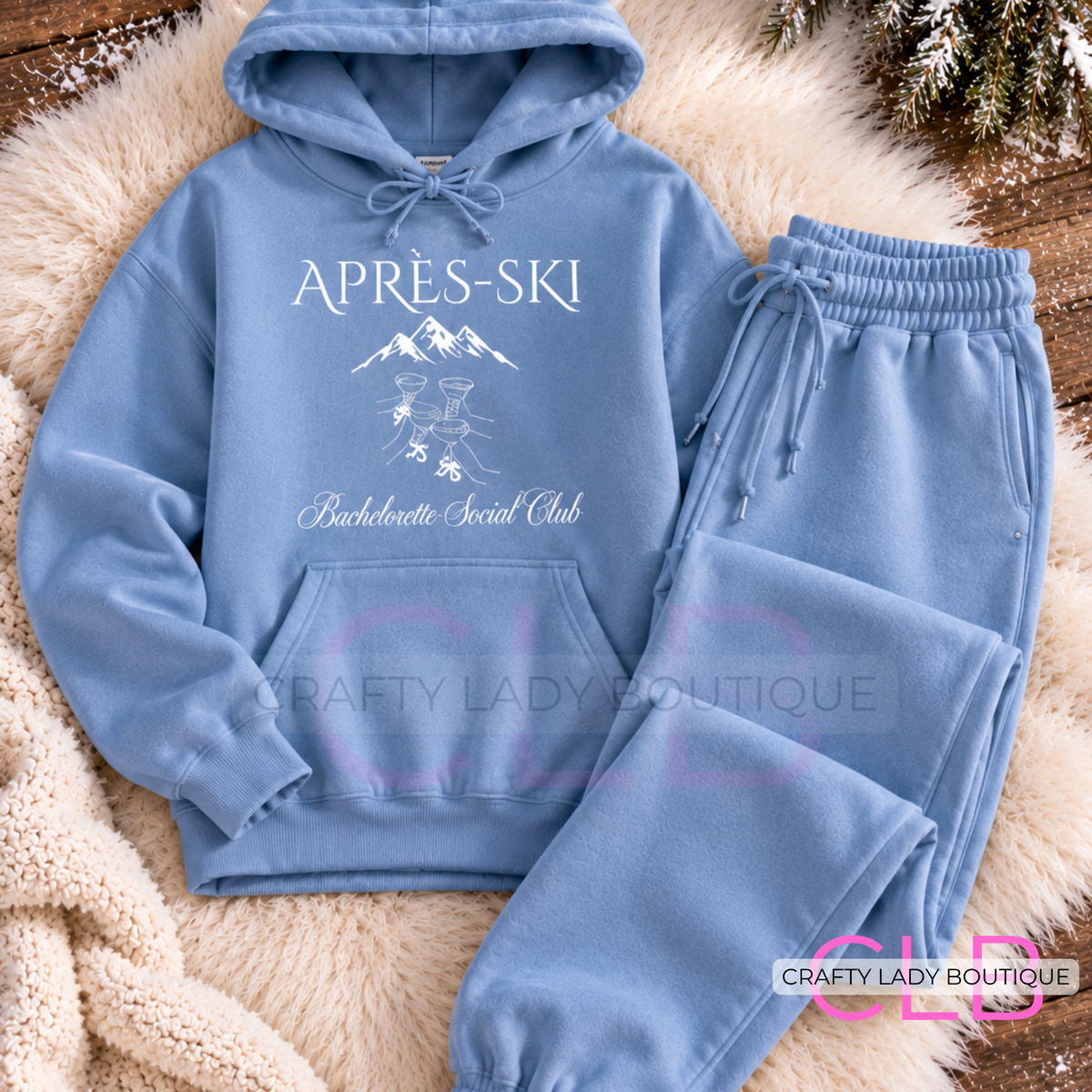 A light blue hoodie and matching sweatpants set laid flat on a cream faux-fur surface with winter foliage at the top. The hoodie has white printed text reading 'APRES-SKI' and 'Bachelorette Social Club' with a small mountain-and-drink graphic.