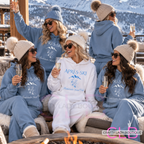 Five women wearing matching ski-themed loungewear sets in light blue and white, sitting around a fire pit with snowy mountains in the background. The sets include hoodies and sweatpants with 'APRÈS-SKI' and mountain graphics printed on the front.