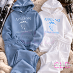 Two matching hooded sweatshirt and jogger set laid flat on a textured beige surface, one in light blue and one in white, each with 'APRÈS-SKI' and mountain graphics printed on the front. Ski poles and skis are partially visible around the edges.