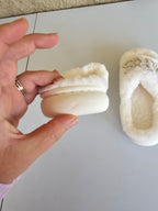 White fuzzy slippers with decorative elements held by a hand against a neutral background
