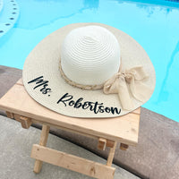 White sun hat with 'Mrs. Robertson' text on a wooden stool by a pool.