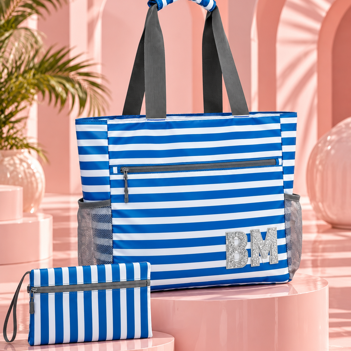 A blue and white striped tote bag with dark gray handles and side mesh pockets, displayed alongside a matching zip pouch on pink display pedestals in a pastel pink studio setting.