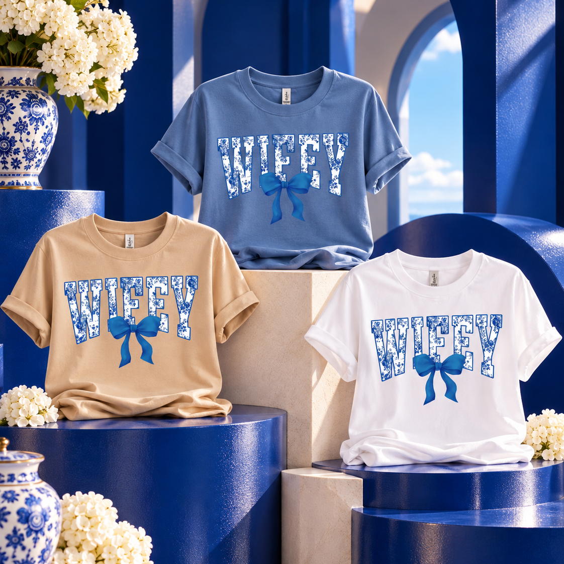 Three graphic t-shirts displayed on blue pedestals: a beige shirt with a tan 'WIFFY' print and blue bow, a light blue shirt with a blue 'WIFFY' print and blue bow, and a white shirt with a blue 'WIFFY' print and blue bow. The background features blue arches, white flowers, and blue-and-white ceramic vases.