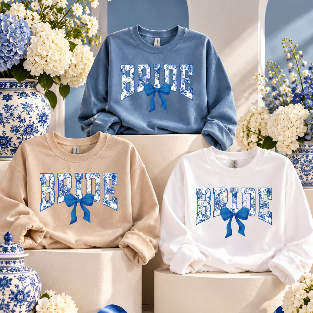 Three crewneck sweatshirts displayed on pedestals in a styled studio setting. The sweatshirts are shown in beige, light blue, and white, each featuring a large 'BRIDE' graphic in a blue-and-white floral pattern with a matching blue bow underneath.