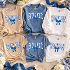 Six crewneck sweatshirts in beige, light blue, and white, each printed with 'BRIDE' in a floral pattern and a blue bow graphic. The sweaters are arranged in a flat-lay with blue ribbons, flowers, and decorative blue-and-white dishes on a light fabric background.