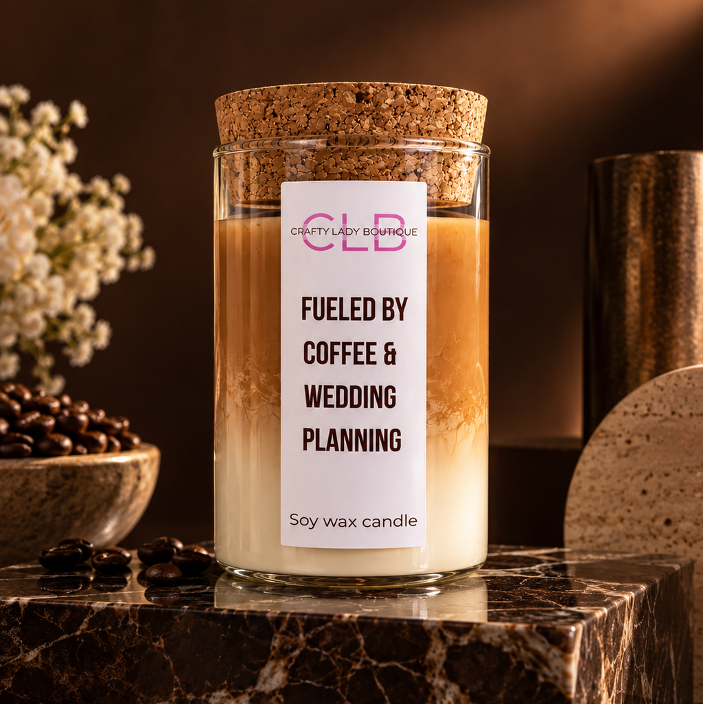 A glass jar candle with a cork lid and a white label reading 'FUELED BY COFFEE & WEDDING PLANNING' and 'CLB Crafty Lady Boutique,' styled on a dark marble surface with coffee beans and warm brown background props.