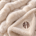 Close-up of a plush cream throw blanket with a chunky braided texture and a folded corner showing a sherpa-like underside with a brown monogram reading AHA.