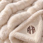 Close-up of a plush cream throw blanket with a chunky braided texture and a folded corner showing a sherpa-like underside with a brown monogram reading AHA.