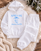 A white two-piece loungewear set consisting of a pullover hoodie and matching sweatpants, laid flat on a beige faux-fur rug over a wooden surface. The hoodie features a blue graphic print with the text 'APRÈS-SKI' and 'Bachelorette Social Club' along with a mountain and skier illustration.