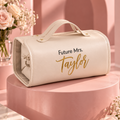 A cream faux-leather cylindrical handbag with a top handle, featuring the personalized text 'Future Mrs. Taylor' in black and gold script, displayed on a pink pedestal with floral props.