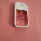 A small white handheld fragrance diffuser bottle with a clear front window showing pink liquid inside, mounted against a solid pink background. The bottle has 'C.