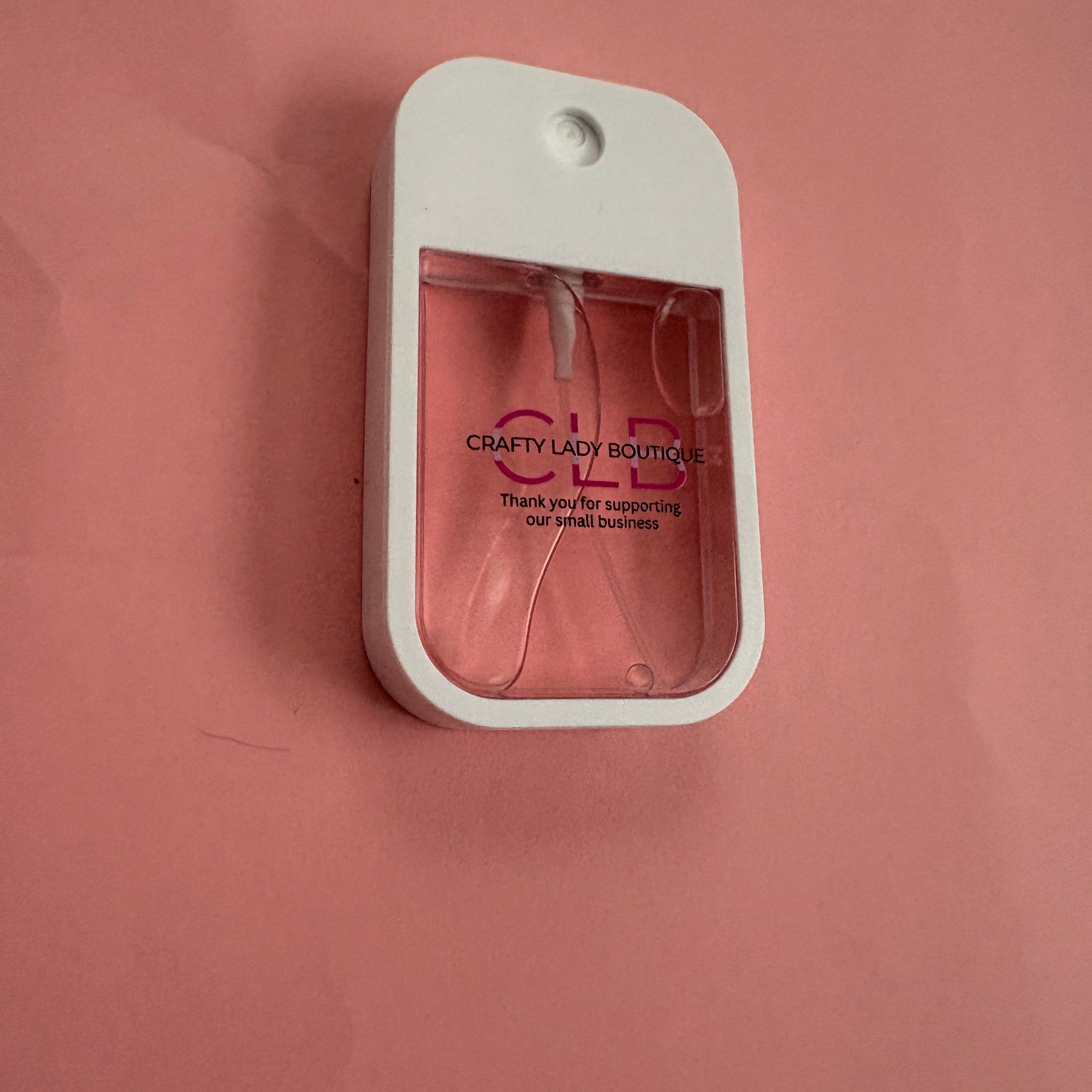 A small white handheld fragrance diffuser bottle with a clear front window showing pink liquid inside, mounted against a solid pink background. The bottle has 'C.