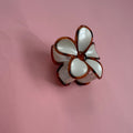 A small flower-shaped accessory with glossy white petals outlined in dark brown, photographed on a plain pink background.