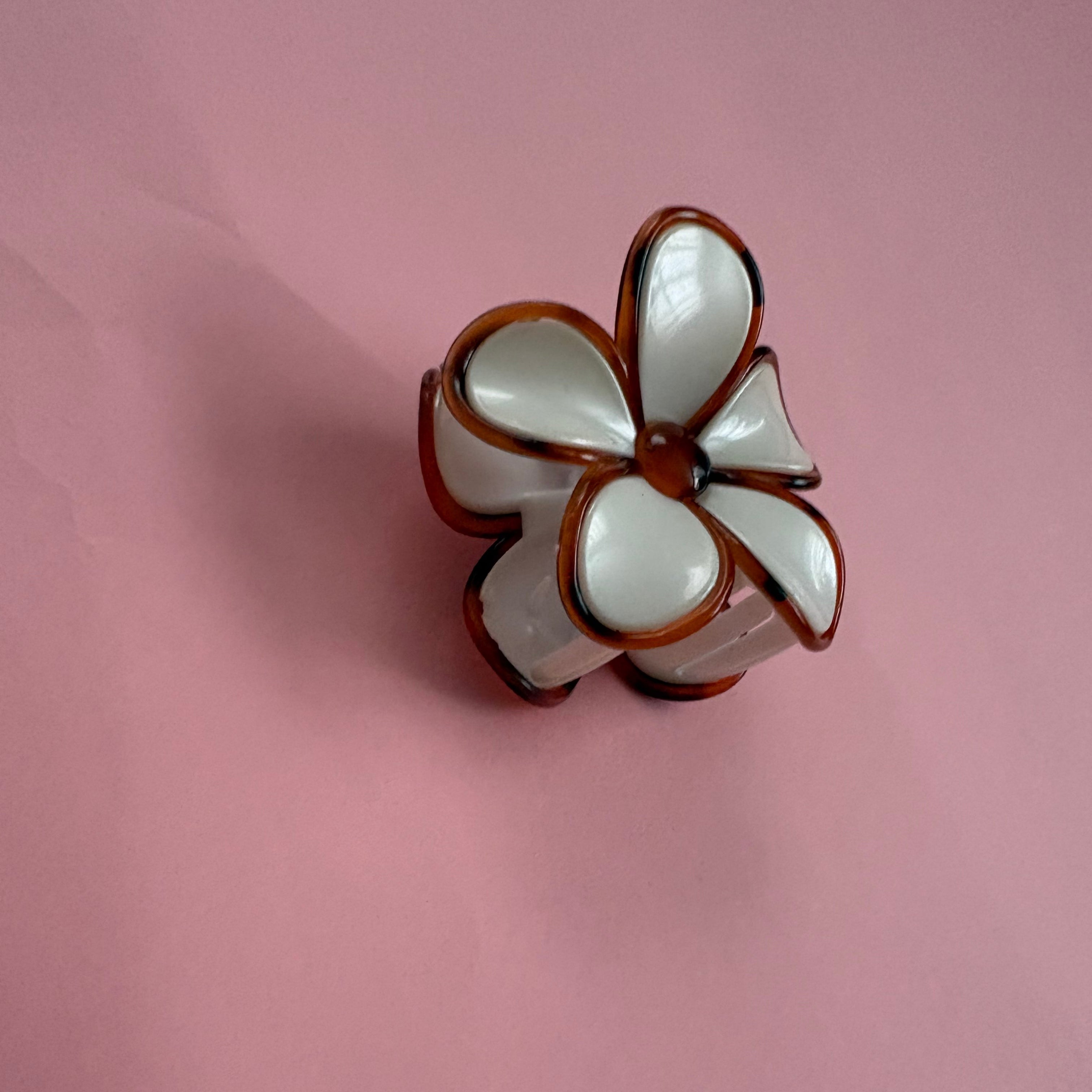 A small flower-shaped accessory with glossy white petals outlined in dark brown, photographed on a plain pink background.