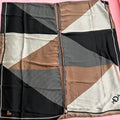 A square scarf laid flat on a pink background, featuring a geometric color-blocked pattern with black, beige, and white sections, including some vertical stripe details and a small black logo in the bottom right corner.