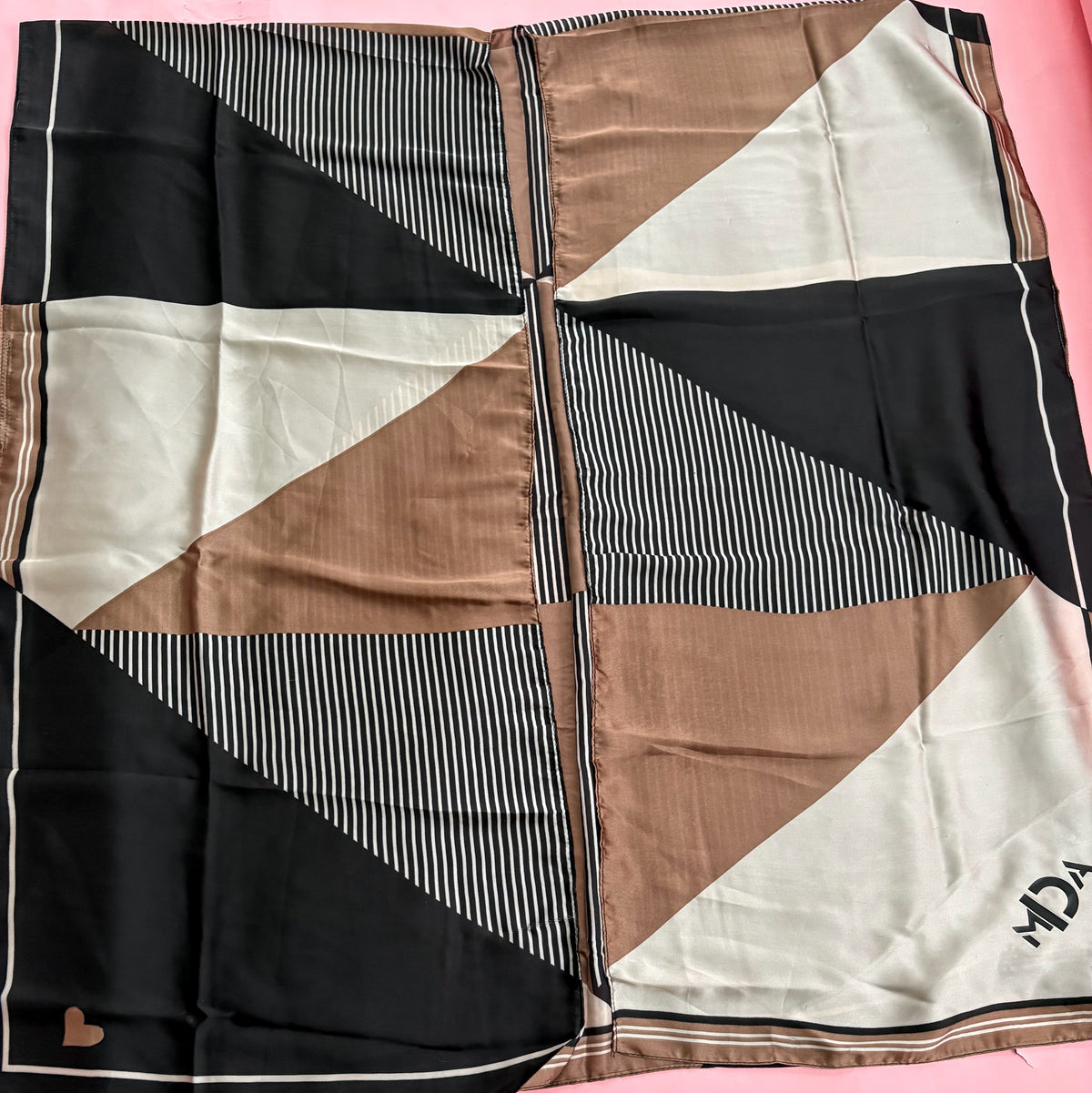 A square scarf laid flat on a pink background, featuring a geometric color-blocked pattern with black, beige, and white sections, including some vertical stripe details and a small black logo in the bottom right corner.