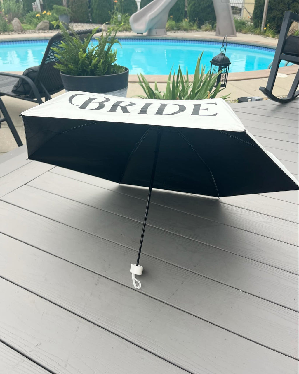 Black umbrella with 'BRIDE' text on a wooden deck near a pool