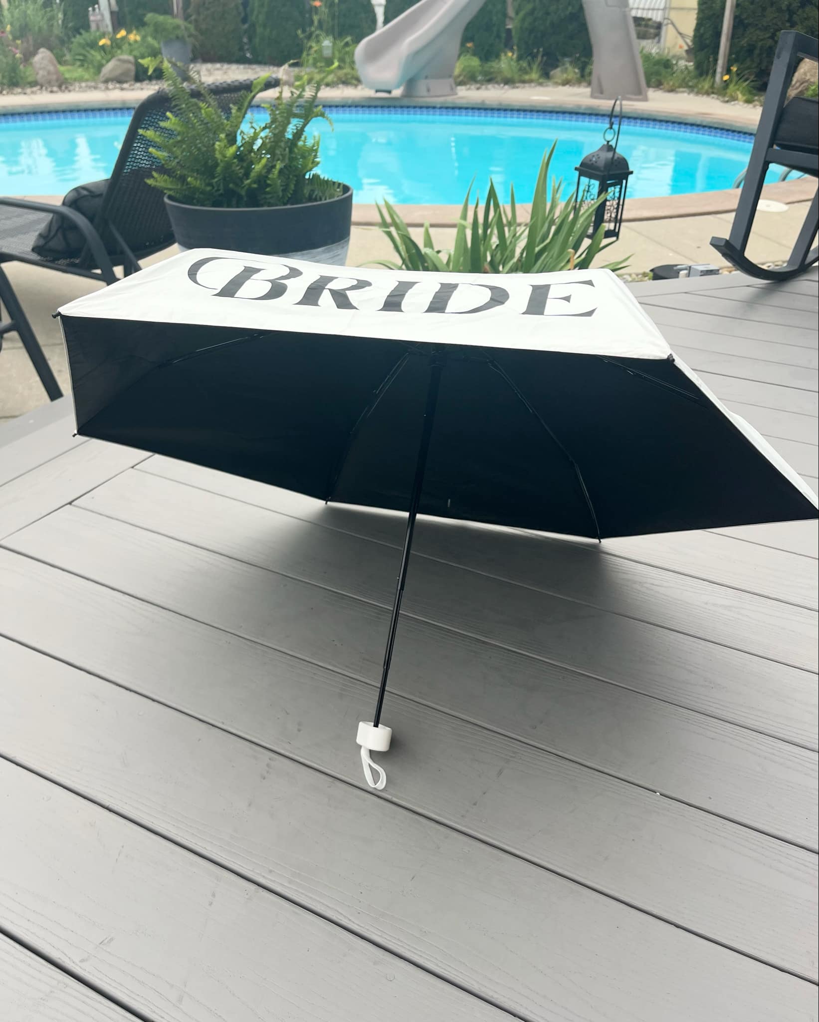 Black umbrella with 'BRIDE' text on a wooden deck near a pool
