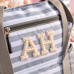 Close-up of a striped tote bag with gray and white horizontal stripes, featuring a top zipper, gray webbing handles, and decorative gold letters embellished with pearls spelling 'AH'.