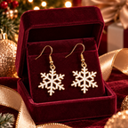 A pair of gold-tone snowflake dangle earrings displayed in an open burgundy velvet jewelry box, surrounded by festive holiday decorations including a Christmas tree, ornaments, and gold ribbons.