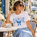 A woman seated at an outdoor cafe wearing a white graphic T-shirt with a blue floral 'BRIDE' design and a light blue satin midi skirt.