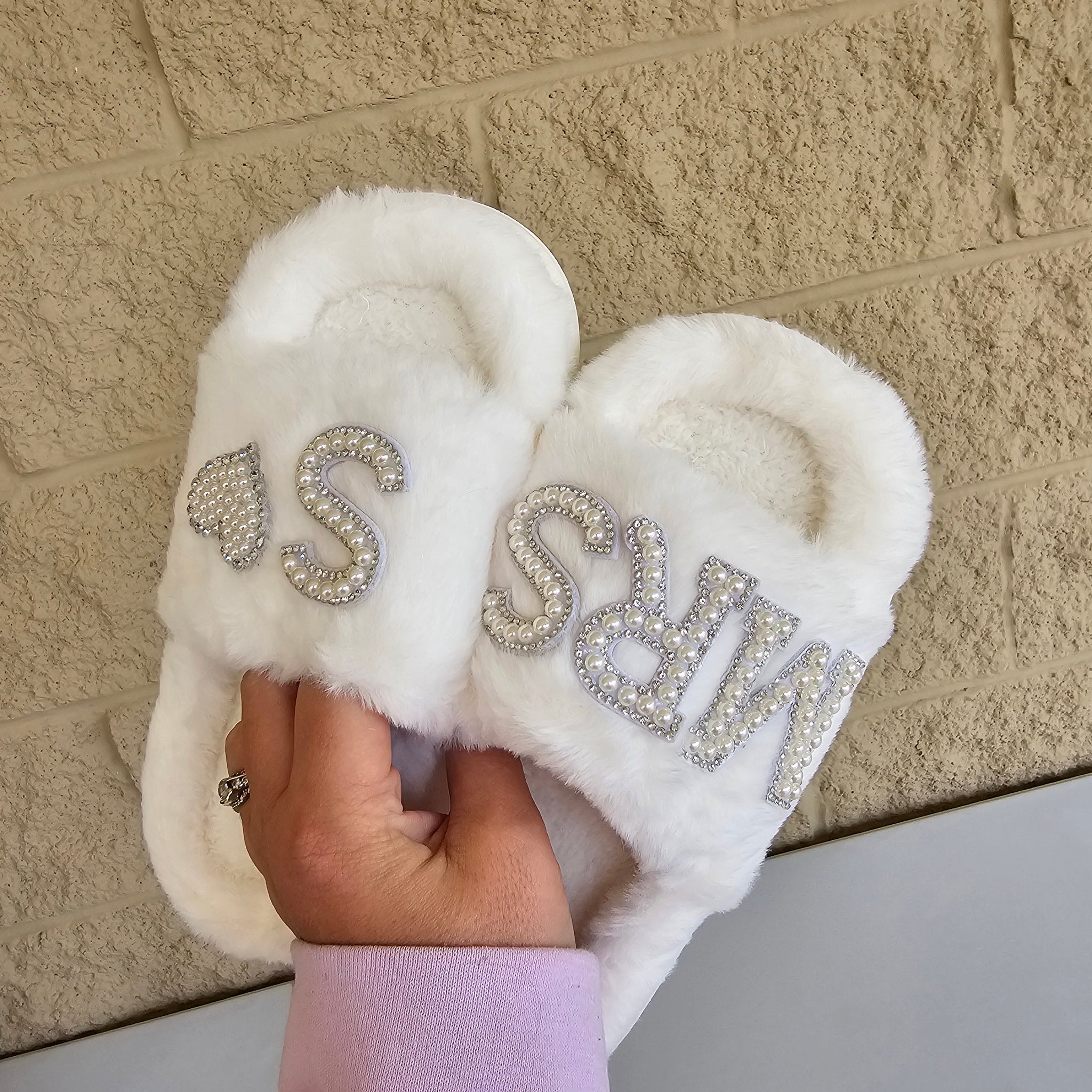 White slippers with rhinestone text held by a hand on a textured surface