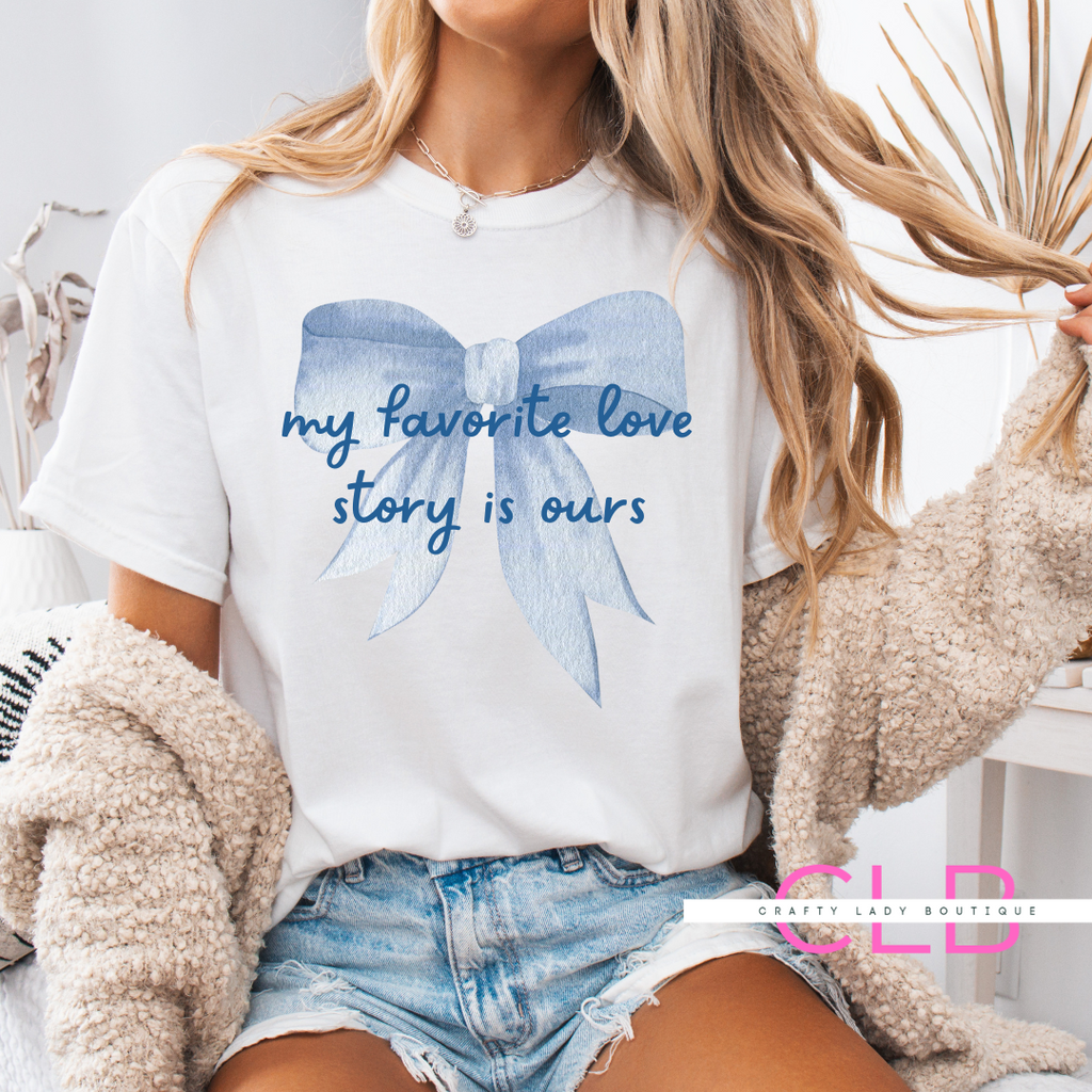 White t-shirt with a large blue bow and the phrase “my favorite love story is ours”—perfect for brides, wives, and romantic occasions.
