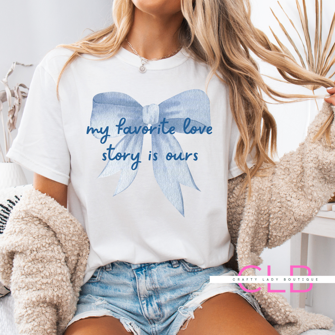 White t-shirt with a large blue bow and the phrase “my favorite love story is ours”—perfect for brides, wives, and romantic occasions.