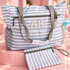 A striped tote bag with gray shoulder straps and a matching zip pouch, displayed on a pink pedestal in a stylized poolside setting. The bag features a light blue and white stripe pattern, gray mesh side pockets, and a monogrammed 'AH' on the front.