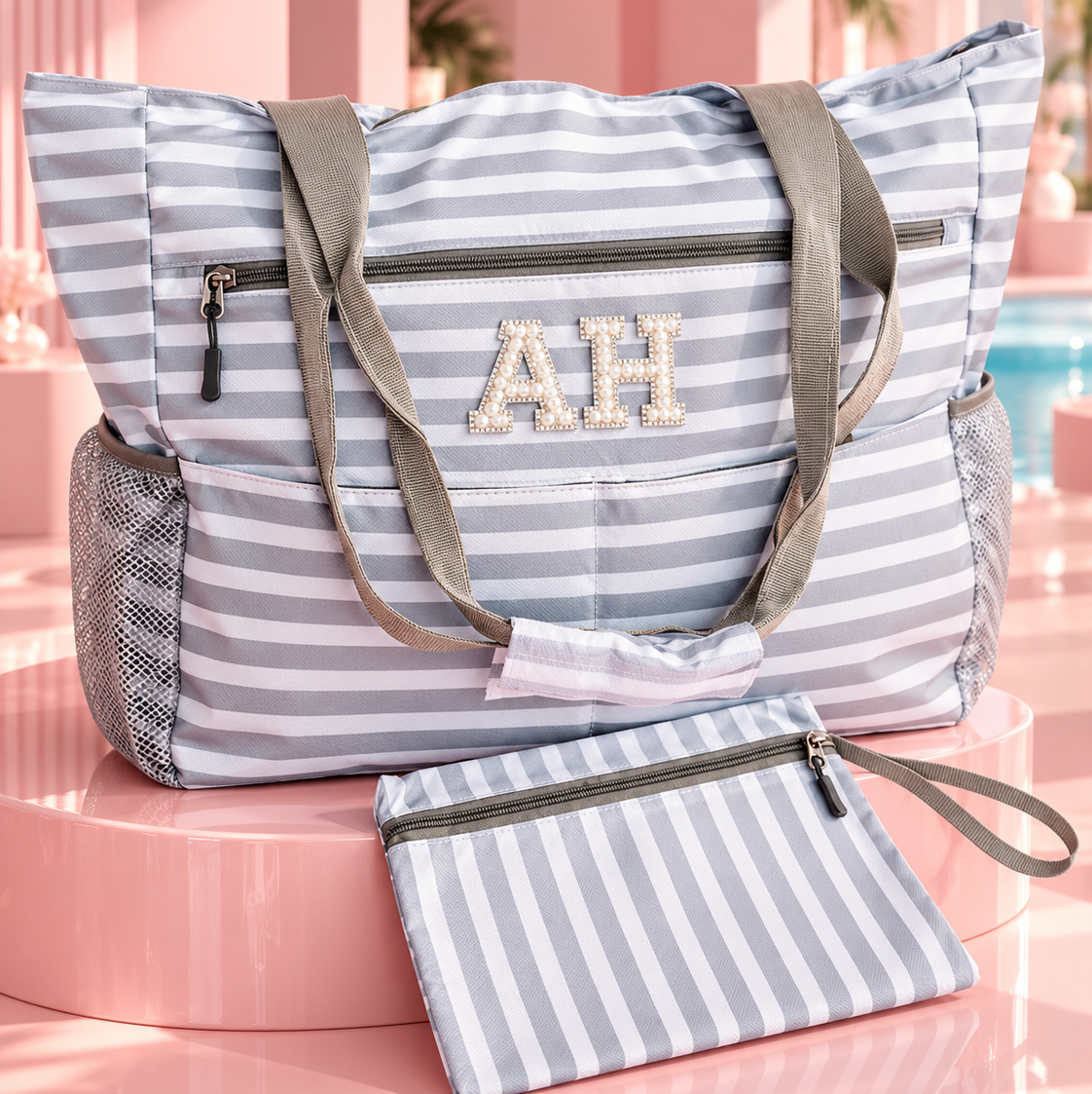 A striped tote bag with gray shoulder straps and a matching zip pouch, displayed on a pink pedestal in a stylized poolside setting. The bag features a light blue and white stripe pattern, gray mesh side pockets, and a monogrammed 'AH' on the front.