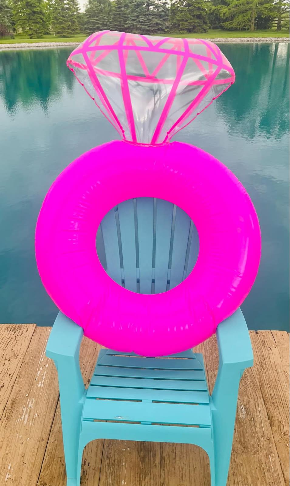 Bright pink inflatable diamond ring pool float displayed on a turquoise chair beside a lake, perfect for brides and bachelorette parties.