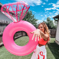 Bright pink inflatable diamond ring pool float displayed on a turquoise chair beside a lake, perfect for brides and bachelorette parties.