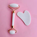 White facial roller and gua sha set with gold hardware displayed on a pink background for skincare and self-care use.