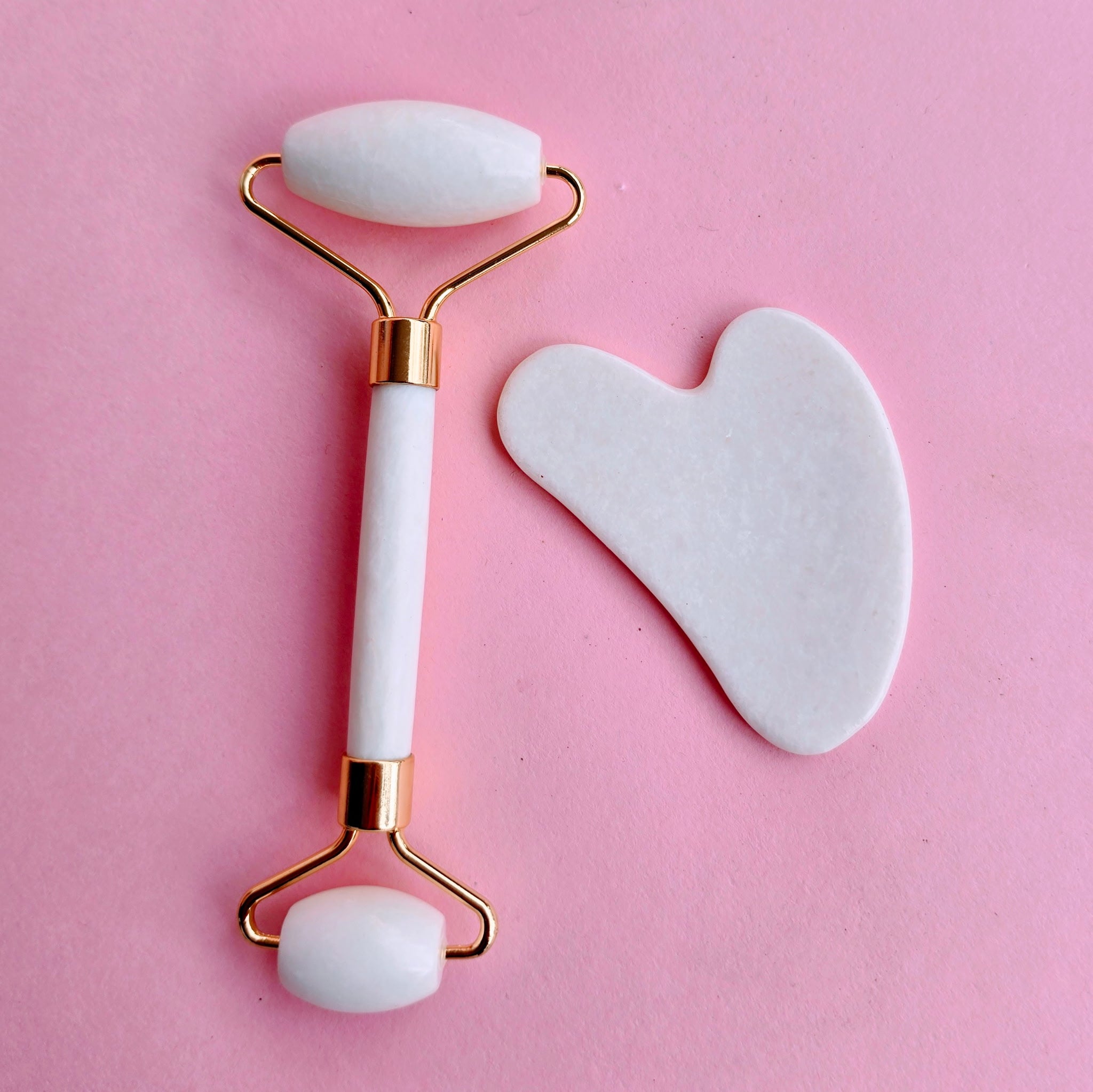 White facial roller and gua sha set with gold hardware displayed on a pink background for skincare and self-care use.