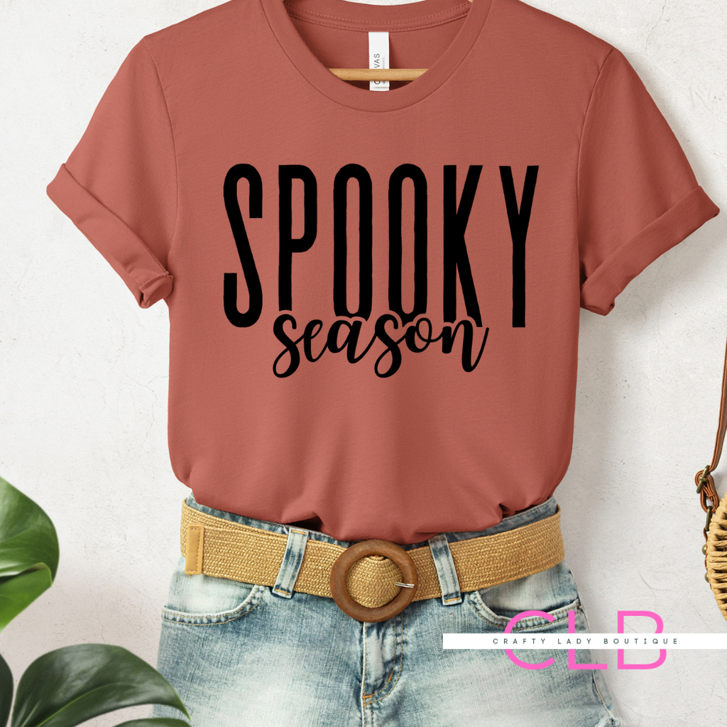 A terracotta-colored short-sleeve t-shirt with 'SPOOKY season' printed in black across the chest, styled on a headless mannequin with a tan woven belt and light-wash denim shorts against a white textured wall.