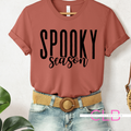 A terracotta-colored short-sleeve t-shirt with 'SPOOKY season' printed in black across the chest, styled on a headless mannequin with a tan woven belt and light-wash denim shorts against a white textured wall.