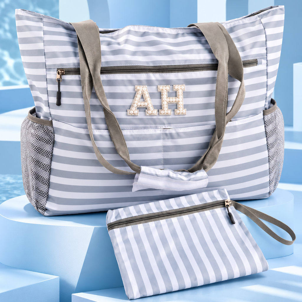 A striped beach tote bag with matching zip pouch, featuring gray and white horizontal stripes, olive green handles, mesh side pockets, and a monogrammed 'AH' on the front, displayed on blue geometric pedestals.