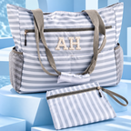 A striped beach tote bag with matching zip pouch, featuring gray and white horizontal stripes, olive green handles, mesh side pockets, and a monogrammed 'AH' on the front, displayed on blue geometric pedestals.