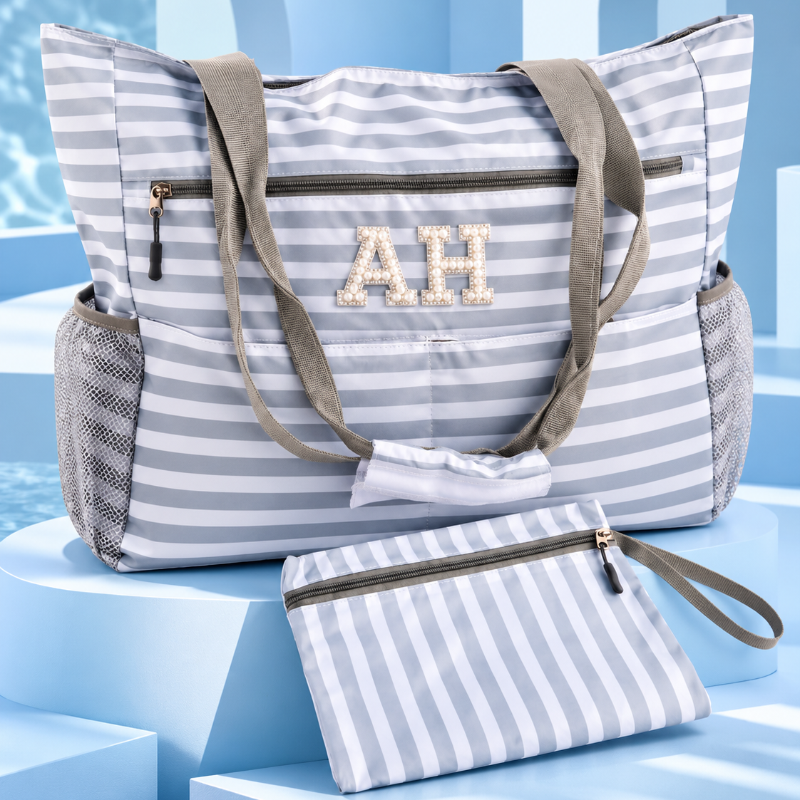 A striped beach tote bag with matching zip pouch, featuring gray and white horizontal stripes, olive green handles, mesh side pockets, and a monogrammed 'AH' on the front, displayed on blue geometric pedestals.