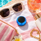 A small blue portable speaker with a black mesh front grille sits on a pink and white striped beach towel next to sunglasses, a drink with orange slices, a gold zipper pouch, a white flower, and a gold chain bracelet.