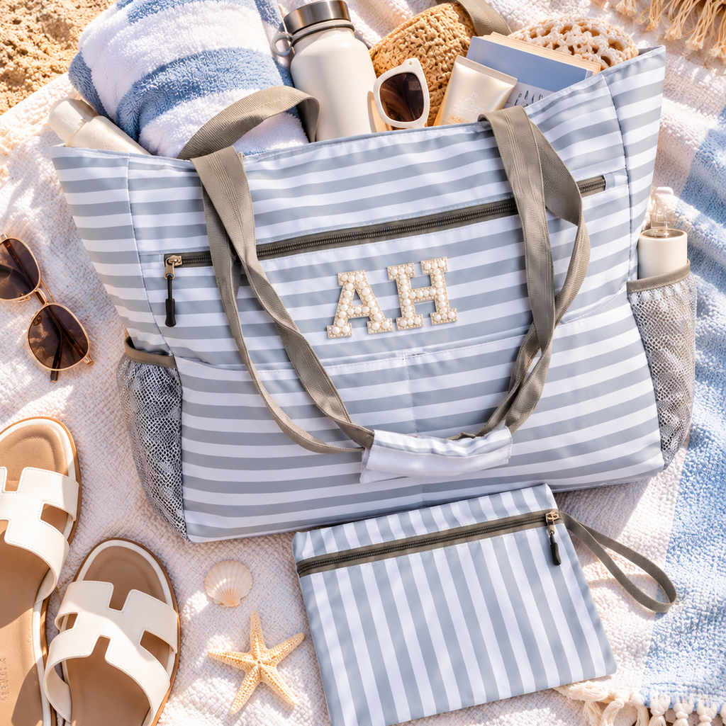 A striped beach tote bag with gray handles and a matching zip pouch, styled on a white blanket with towels, sandals, sunglasses, and beach accessories. The tote has a monogram 'AH' on the front.