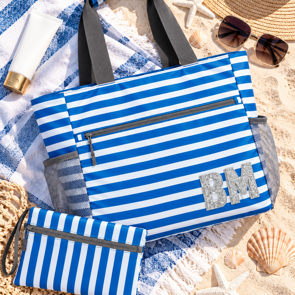 A blue and white striped beach tote bag with gray handles and a matching zip pouch, styled on a sandy beach with a striped towel, sunglasses, a straw hat, and starfish.