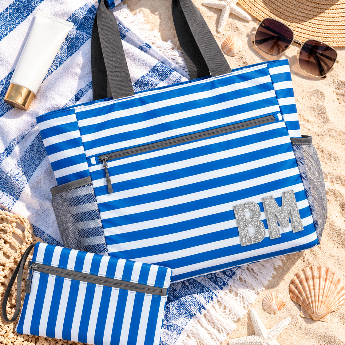 A blue and white striped beach tote bag with gray handles and a matching zip pouch, styled on a sandy beach with a striped towel, sunglasses, a straw hat, and starfish.