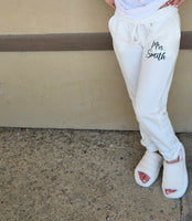 Person wearing white sweatpants with 'Mrs. Smith' printed on the side, standing against a beige wall.