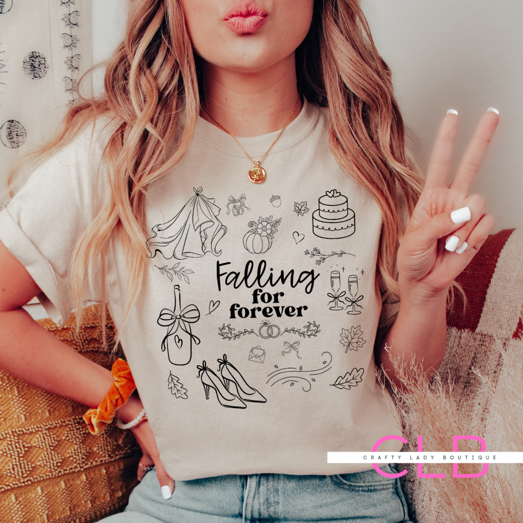 A woman wearing a cream-colored graphic t-shirt with black line drawings of a wedding dress, cake, champagne, and high heels, with the text 'Falling for forever'. She is posing indoors with a peace sign, wearing a gold necklace and blue jeans.