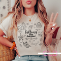 A woman wearing a cream-colored graphic t-shirt with black line drawings of a wedding dress, cake, champagne, and high heels, with the text 'Falling for forever'. She is posing indoors with a peace sign, wearing a gold necklace and blue jeans.