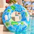 A large inflatable pool float with a tropical blue and green palm leaf pattern, resting on the edge of a swimming pool next to beige stone steps and resort lounge chairs.